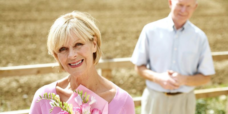 smiling woman holding pink posey and husband in background with questioning pose